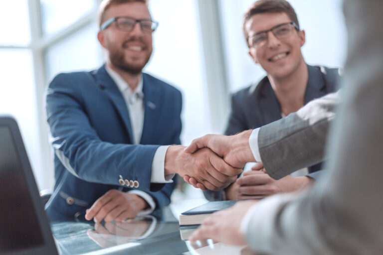 Two businessmen in suits shake hands across a glass table in a bright office setting.