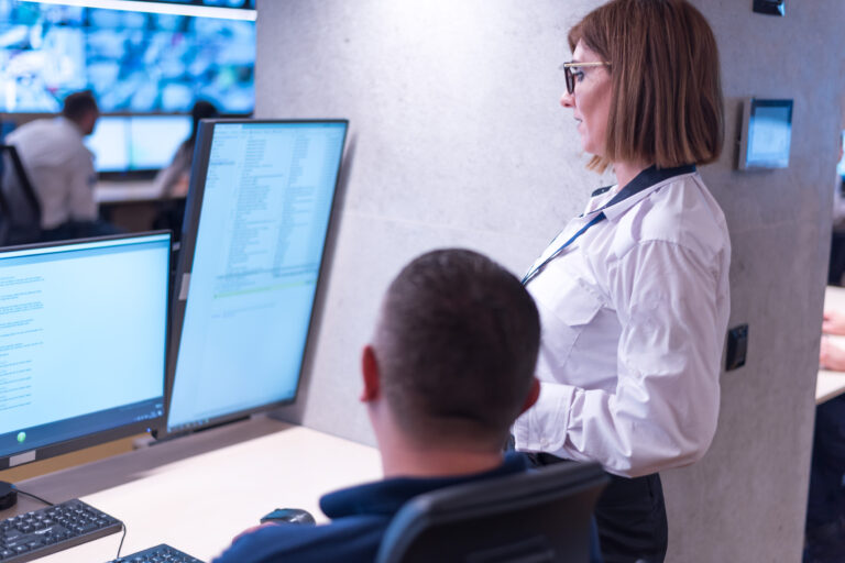Rear view of a female supervisor standing over a male security operator's shoulder, guiding him through data on computer screens in a monitoring room.